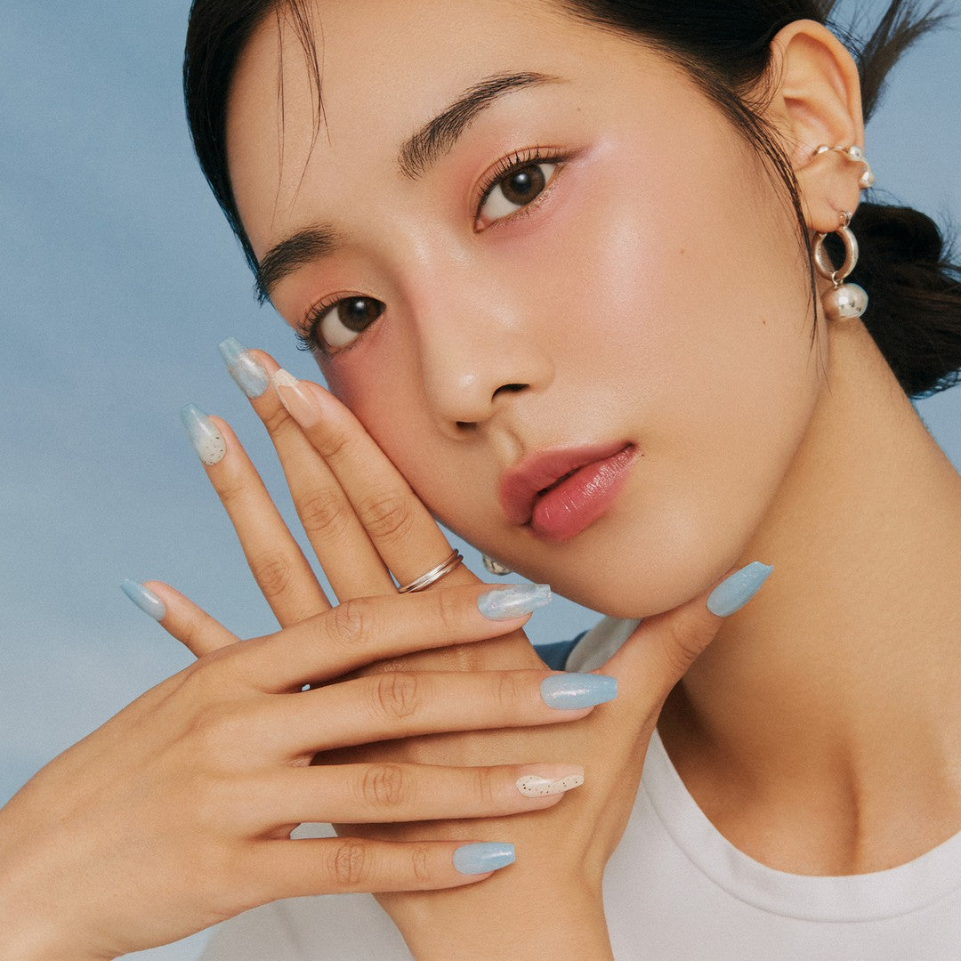 Woman with elegant blue nail art poses with hands near face against blue background, showcasing trendy beauty and style.
