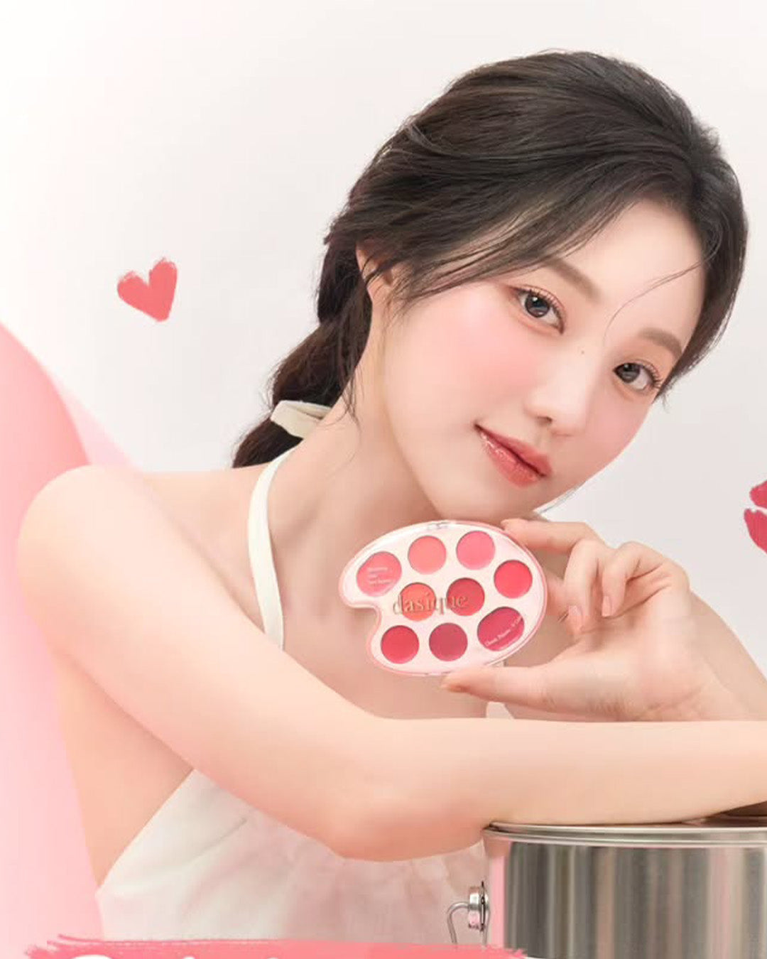 Woman posing with a pink makeup palette, surrounded by hearts, highlighting beauty and cosmetics.