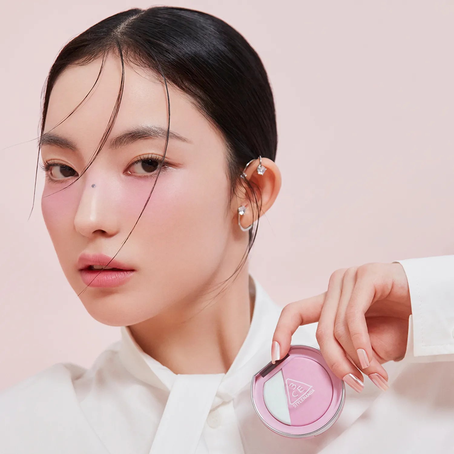 Model displaying 3CE Blushlighter on cheeks with product in hand, demonstrating vibrant and stylish makeup look.