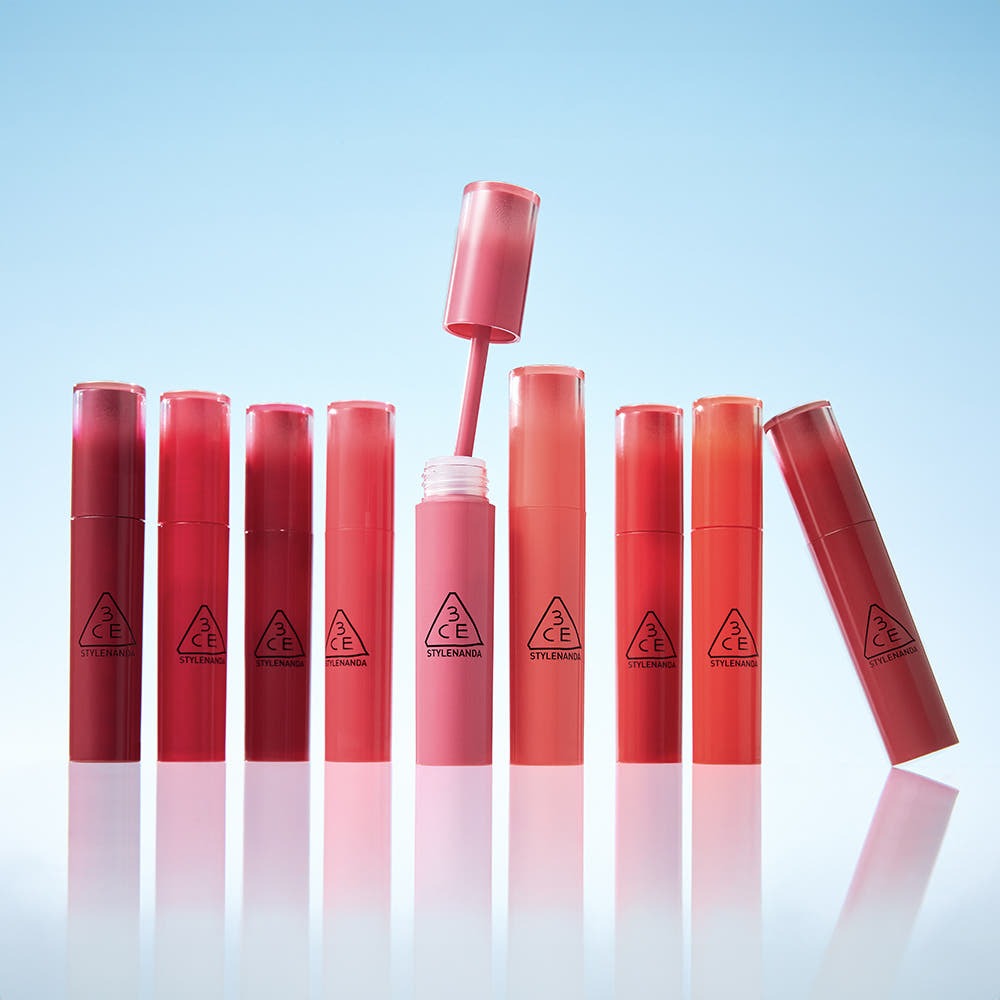 Eight tubes of 3CE Lazy Pop Lip Stain arranged on a reflective surface against a blue background, showcasing vibrant and diverse lip colors.