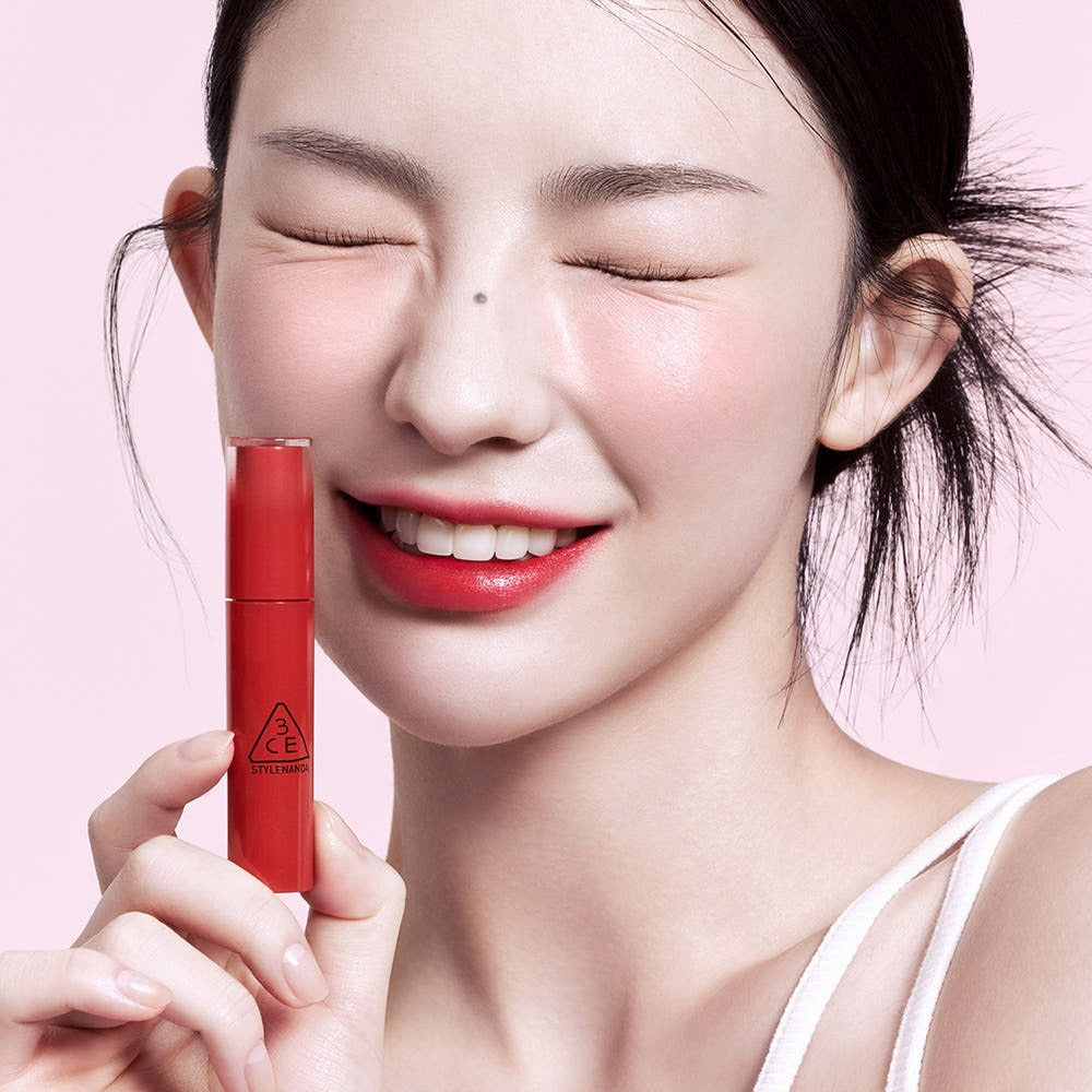 Woman smiling and holding 3CE Lazy Pop Lip Stain in red shade.