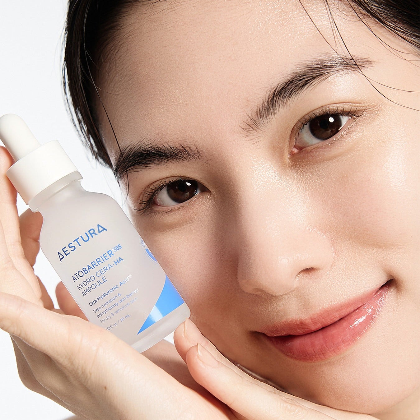 Smiling person holding AESTURA Atobarrier 365 Hydro Cera-HA Ampoule 30ml near face, showcasing the skincare product's hydrating benefits.