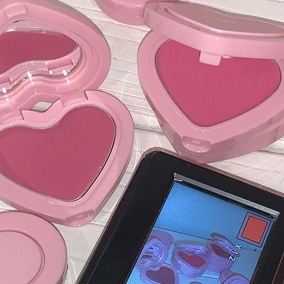 Heart-shaped pink AOU Soft Cream Blush in open compact cases next to a digital display on a wooden surface.