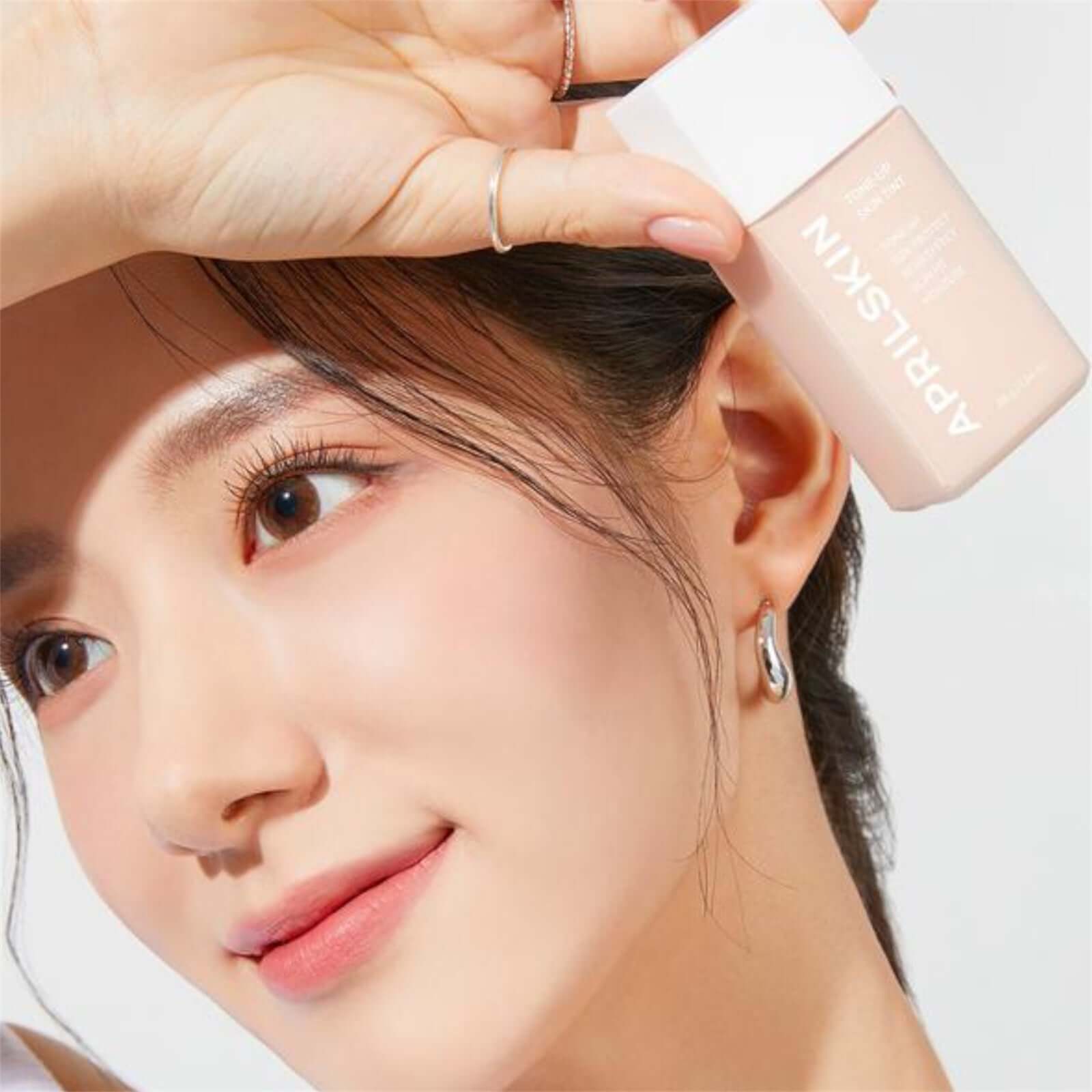 Woman holding APRILSKIN Tone-Up Skin Tint showcasing glowing skin and makeup application.