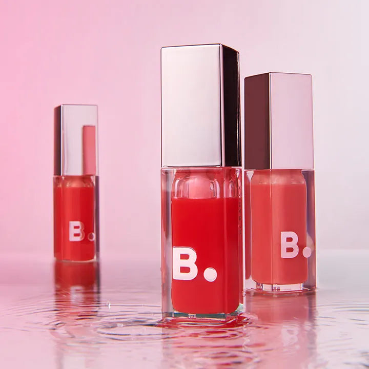 BANILA CO Hydrating Lip Oil bottles on a reflective surface with a pink background.