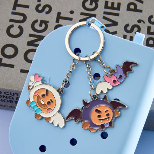 Porte-clés métallique BT21 Angel & Villain avec charms d'ange et de méchant avec une boîte bleue en arrière-plan.