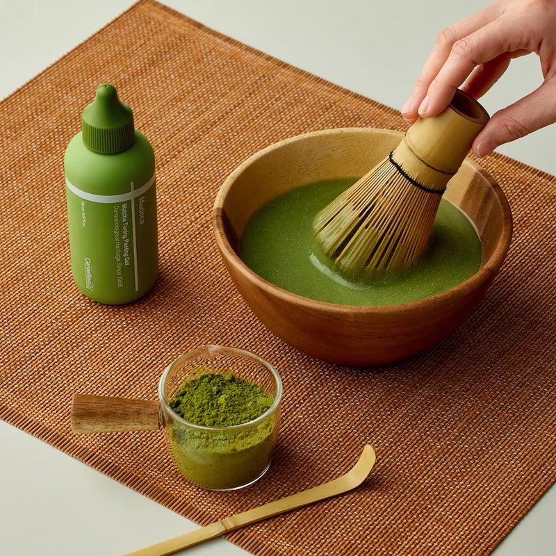 Matcha tea preparation with a bamboo whisk, cup of matcha powder, and a green bottle on a brown mat. Ideal for matcha enthusiasts.