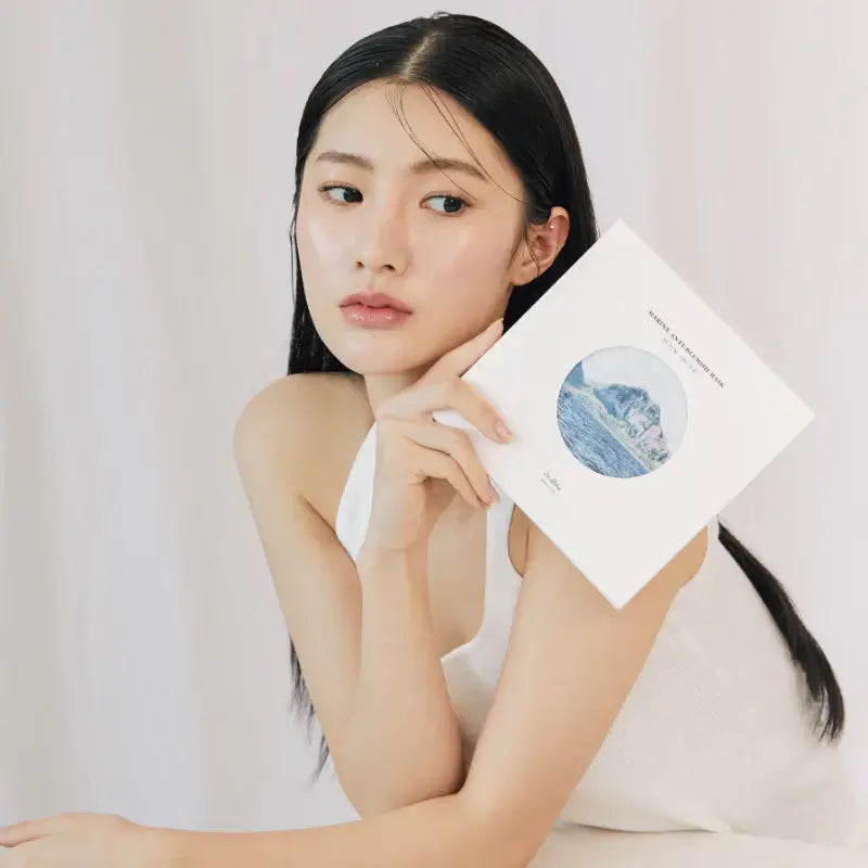 Woman holding DR.ALTHEA Marine Anti Blemish Mask (5ea) in hand wearing a white top with a neutral expression.