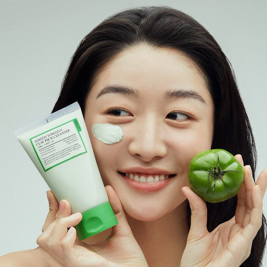 Woman holding FULLY Green Tomato Clay Pack Cleanser 120ml with a green tomato, smiling with clay on cheek.
