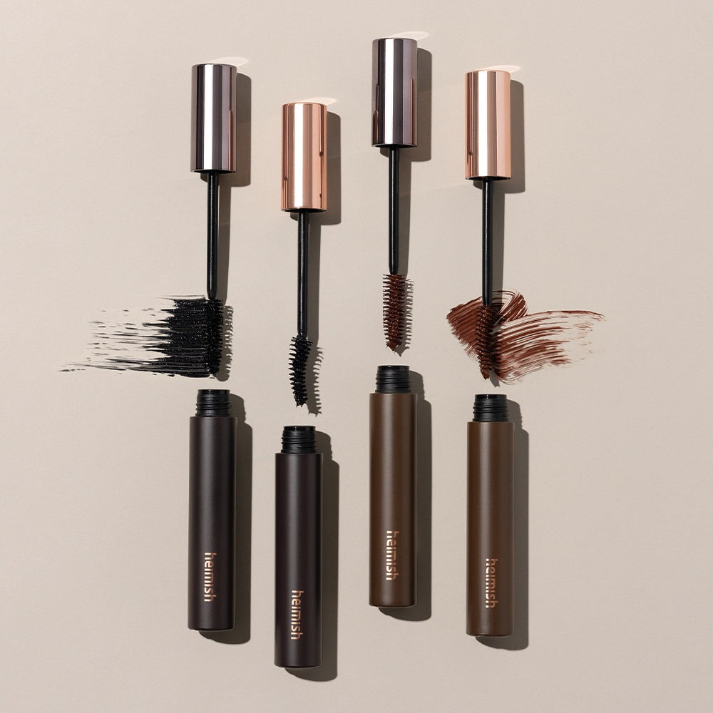 HEIMISH Dailism Mascara in black, brown, and dark brown shades, showing open tubes and brush strokes on a beige background.