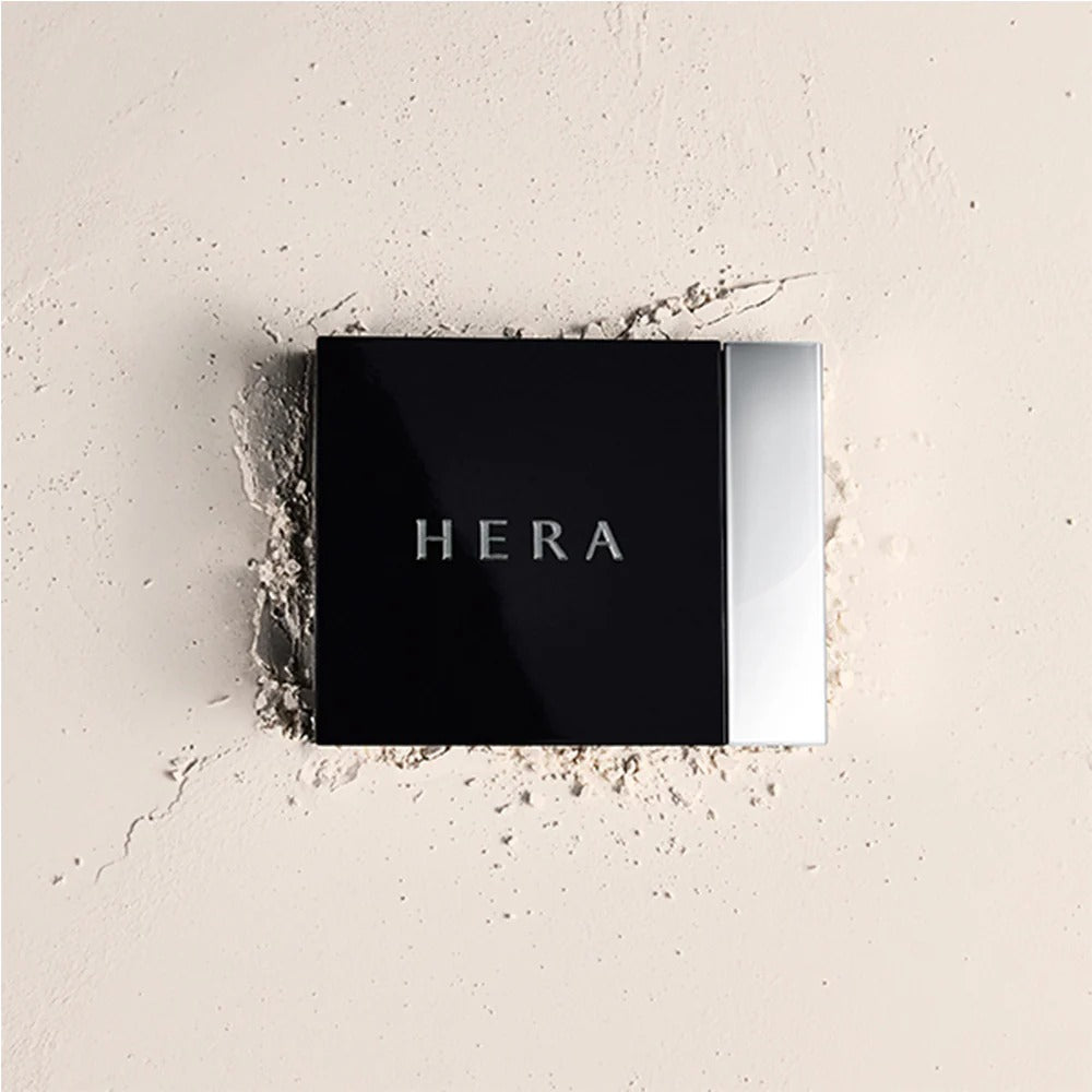 Close-up of HERA Airy Powder Primer 8.5g on a textured surface, showcasing sleek packaging and brand name.