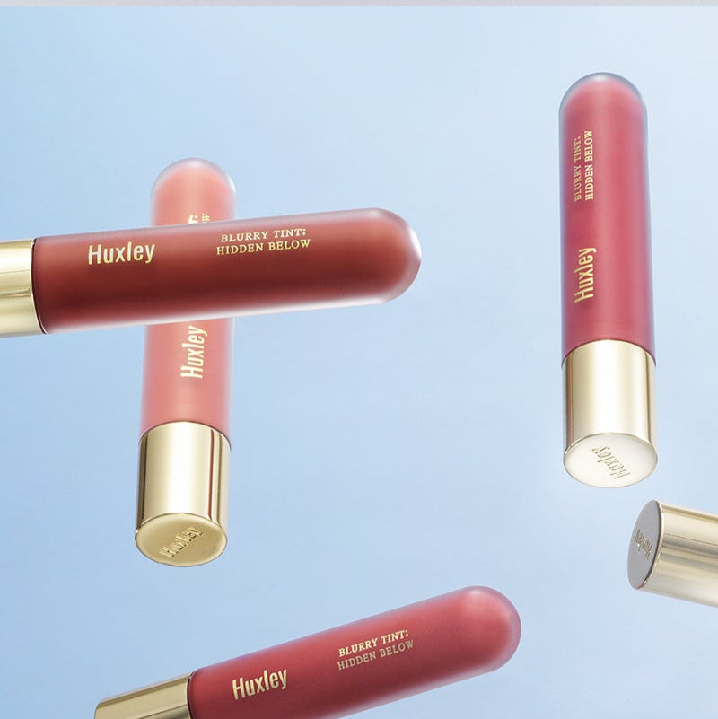 HUXLEY Blurry Tint Hidden Below cosmetic tubes floating in front of a blue background, showcasing elegant design and color.