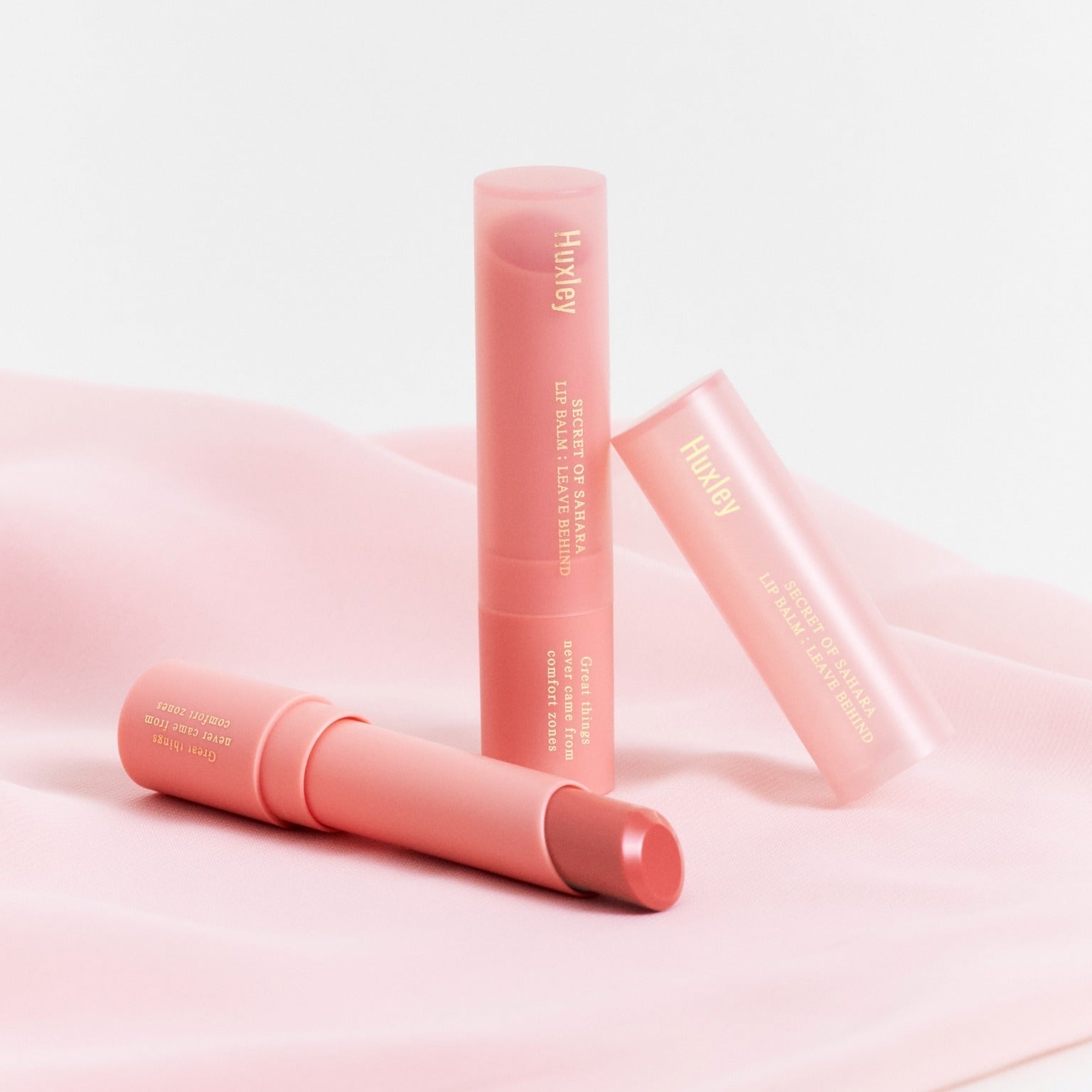 HUXLEY Lip Balm Leave Behide on pink fabric backdrop, showcasing three tubes of the product.