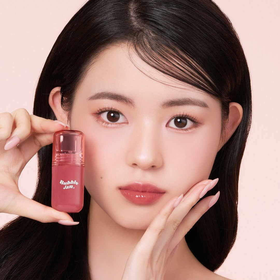 Woman holding IPKN Bubble Jam Gloss, highlighting lip gloss packaging and color on a soft pink background.