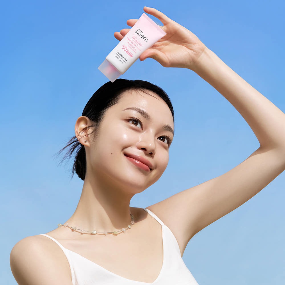 Smiling person holding MAKE P:REM Soothing Pink Tone-Up Sunscreen SPF 50+ PA++++ 50ml against a clear blue sky.
