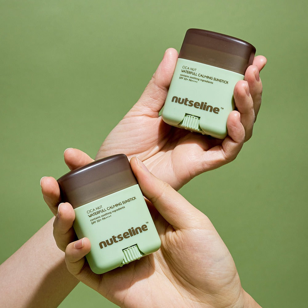 Two hands holding NUTSELINE Cica Nut Waterful Calming Sun Stick 20g against a green background.
