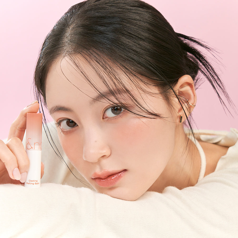 Woman holding ROMAND Glasting Melting Balm (#01 to #15) with smooth complexion, showcasing product's moisturizing effect on lips.