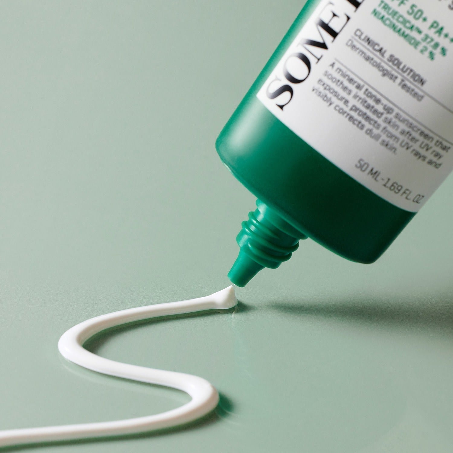 SOME BY MI Truecica Weaponless Calming Tone Up Sun Cream 50ml being dispensed from tube, showcasing texture and color on green surface.