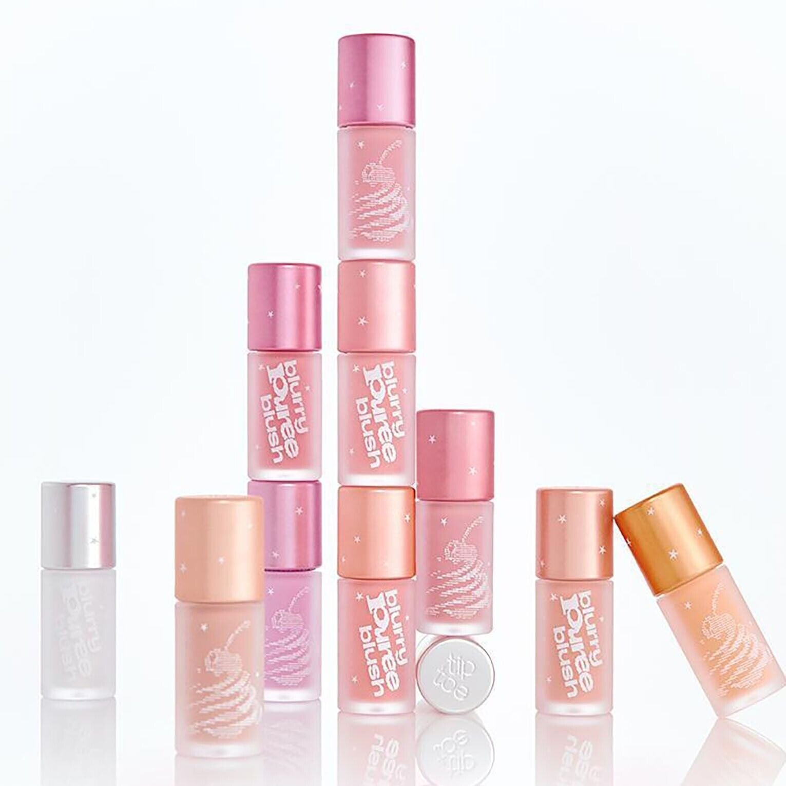Assorted shades of TIP TOE Blurry Puree Blusher bottles stacked creatively against a white background, showcasing packaging design.