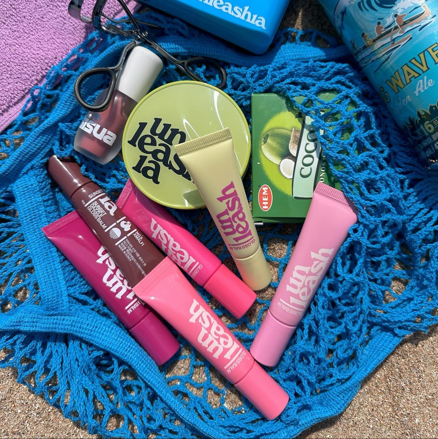 Various UNLEASHIA products including the UNLEASHIA Sunset Dazzle Gloss Balm displayed on a blue net.