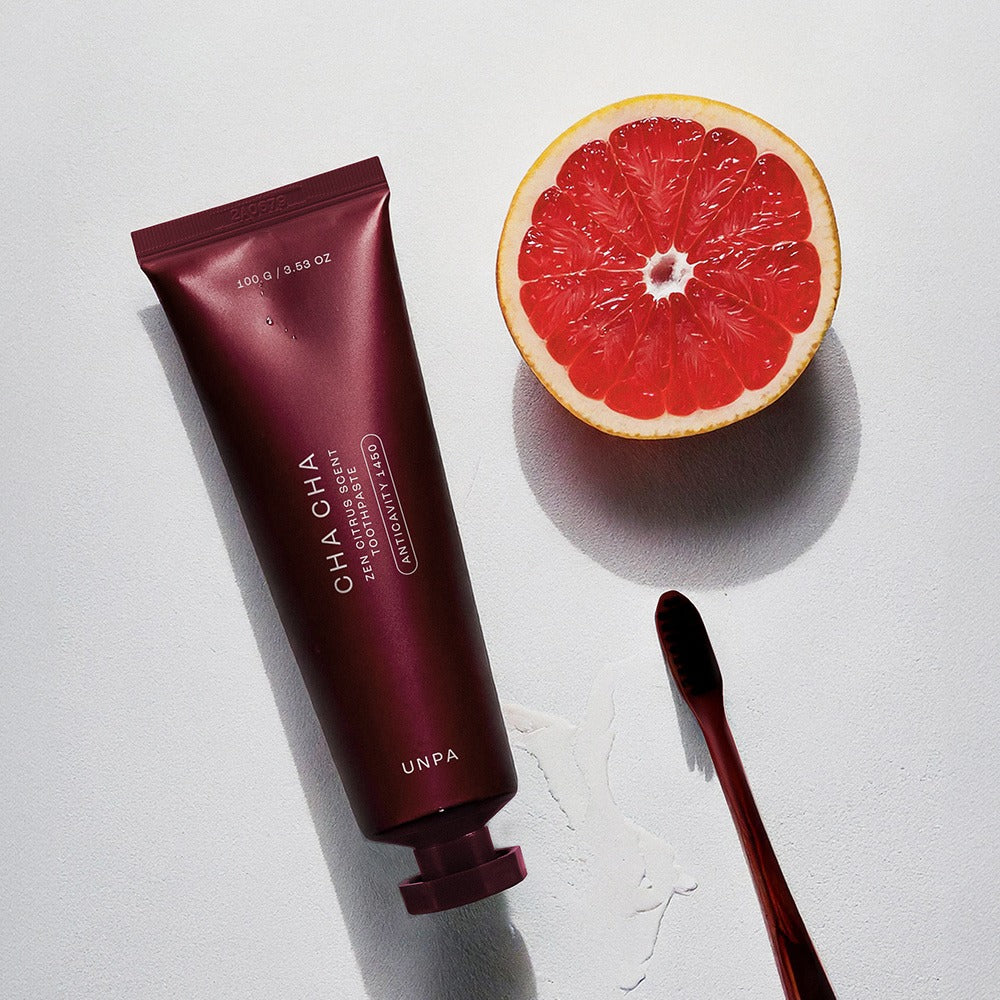 UNPA Cha Cha Zen Grapfruit Scent 1450 High Fluoride Toothpaste 100g with fresh grapefruit half and sleek toothbrush on surface.