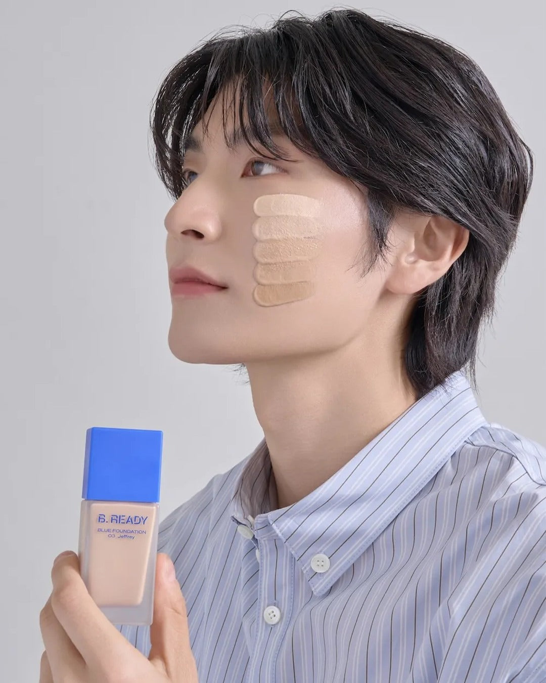 Person holding B-READY foundation bottle with swatches on cheek, demonstrating product shade options.