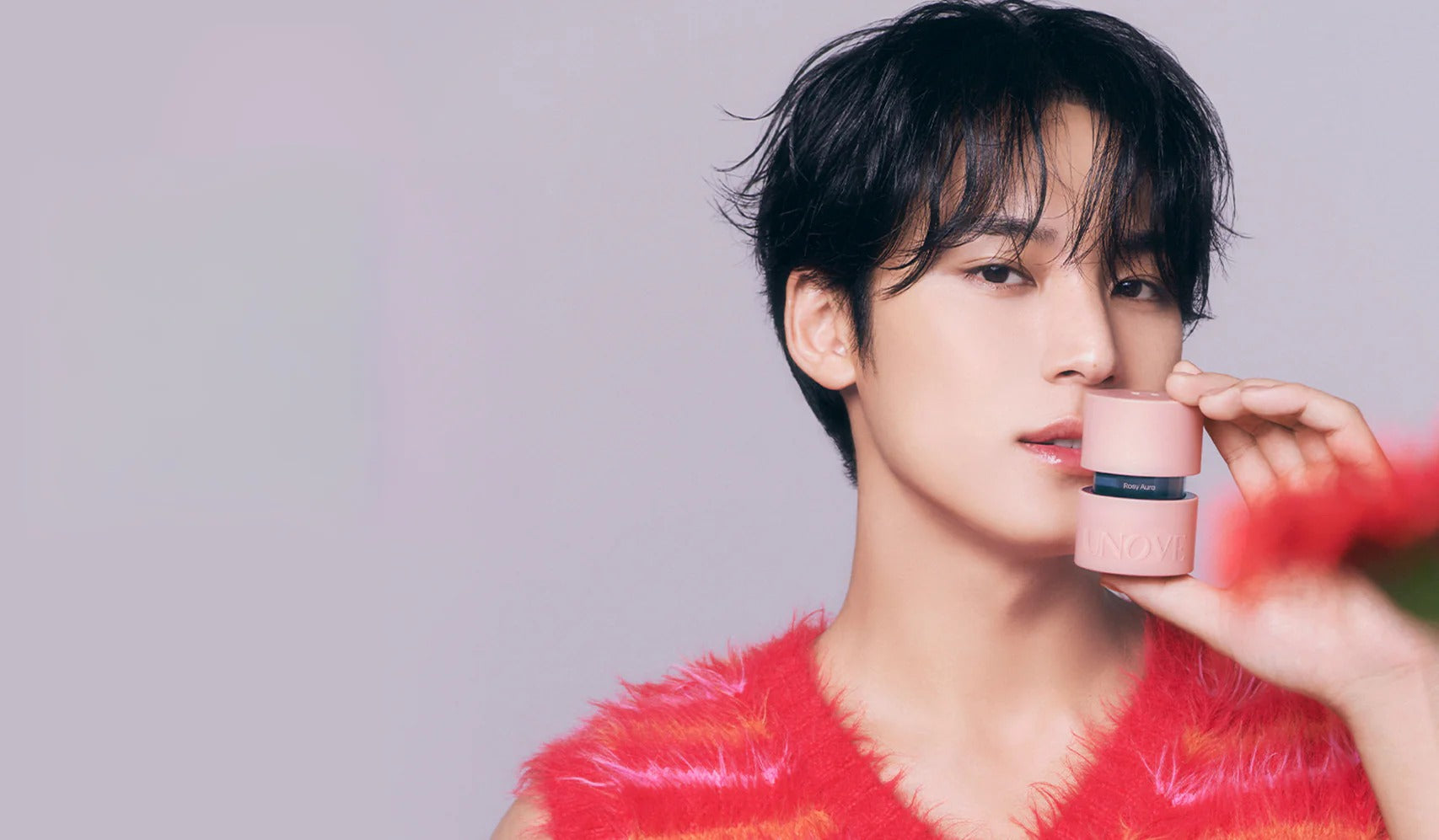 Person posing with skincare product in pink container, wearing a red textured top, against a light background.