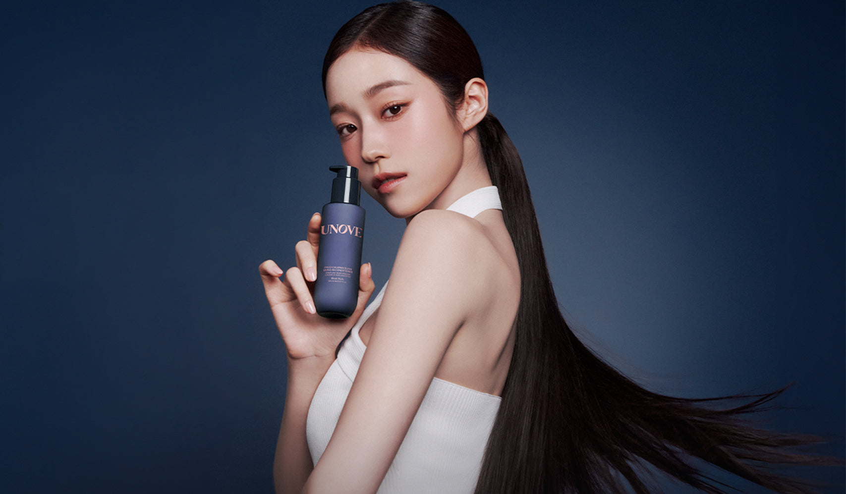 Model holding beauty product bottle against blue background with sleek hairstyle.