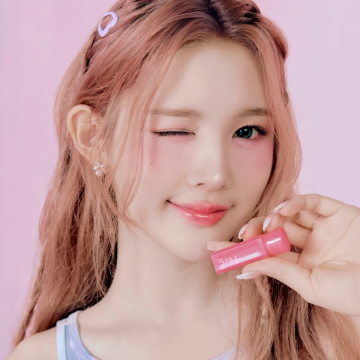 Woman holding 2an Mini Glaze Bouncing Tint in pink packaging, smiling with a wink against a pastel pink background.