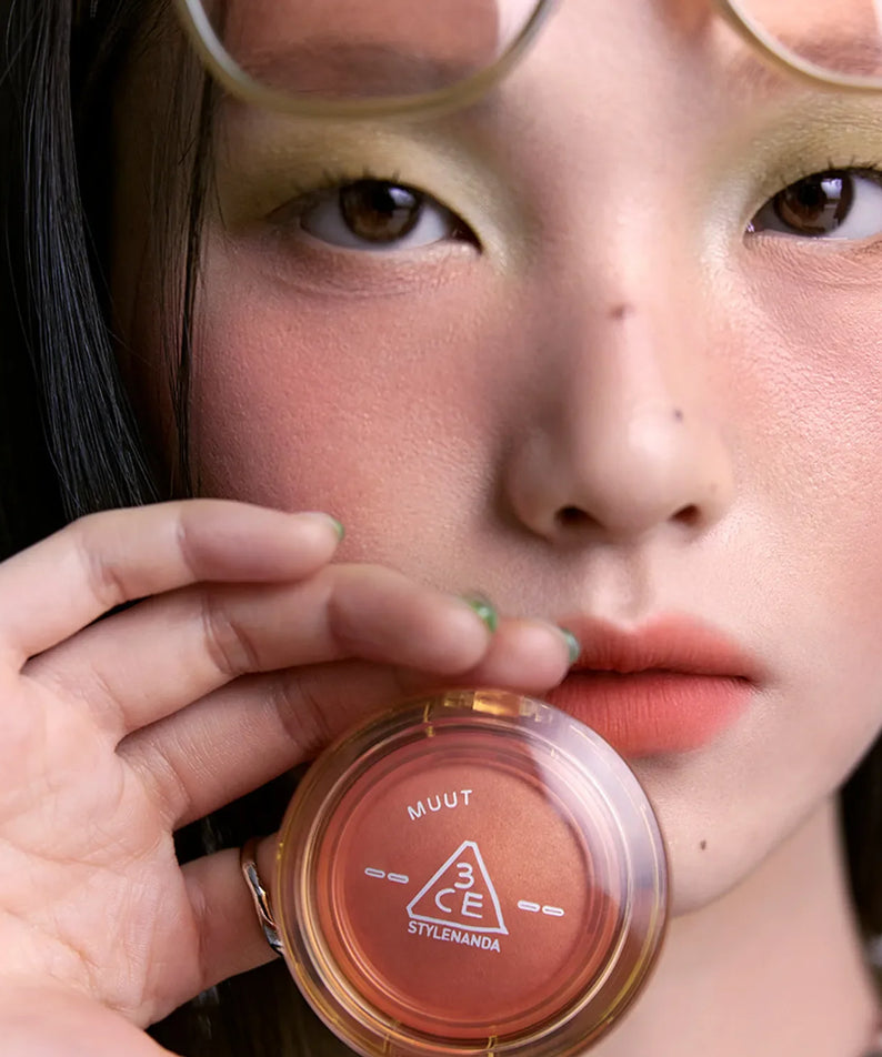 Close-up of a model holding 3CE Bouncy Blur Balm, showcasing its compact design and vibrant color, ideal for a flawless makeup finish.