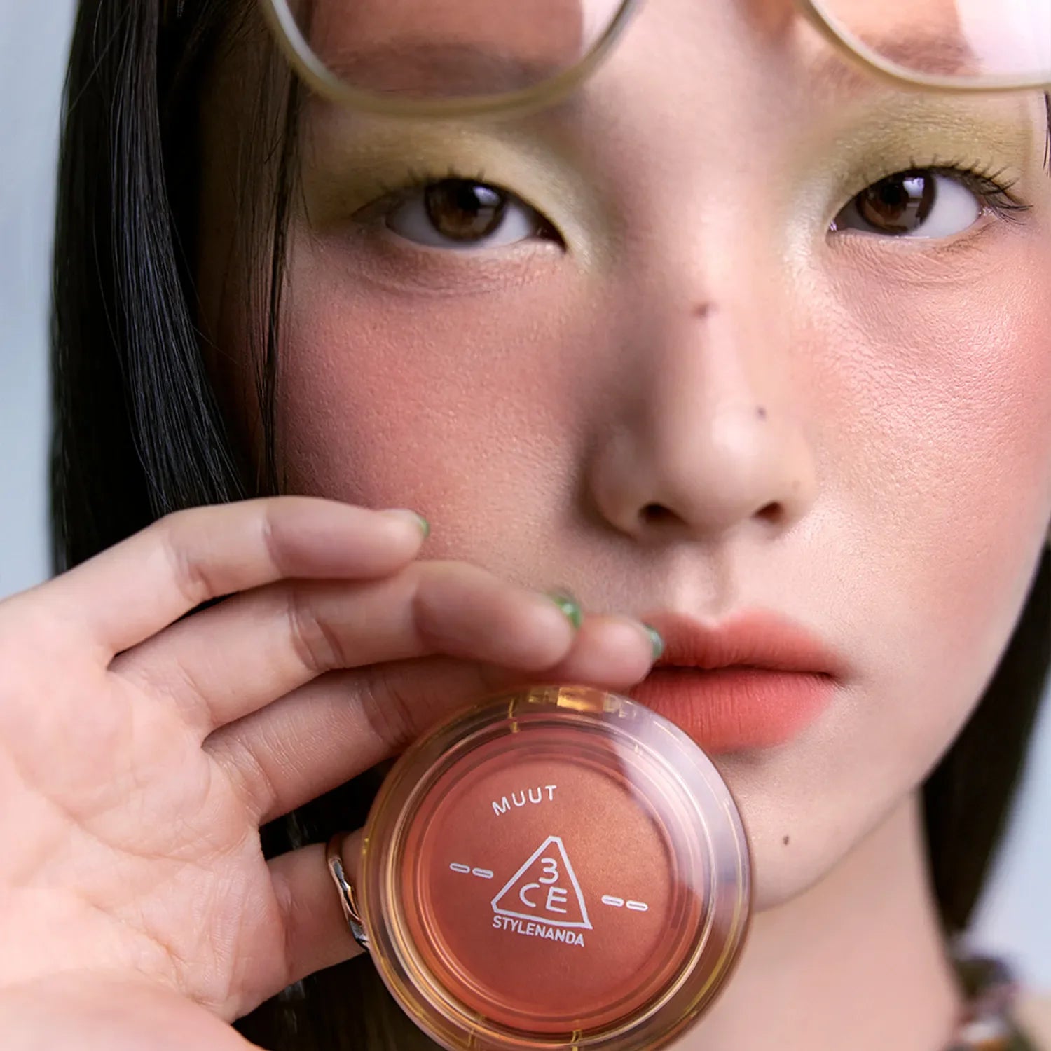 Close-up of a model holding 3CE Bouncy Blur Balm, showcasing its compact design and vibrant color, ideal for a flawless makeup finish.