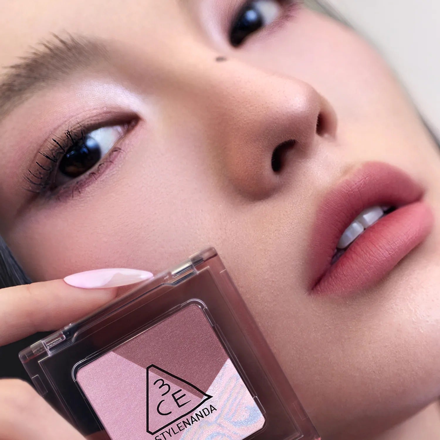 Close-up of woman holding 3CE Color Grid Eyeshadow in pink shade, showcasing subtle eye makeup and soft pink lips for a natural look.