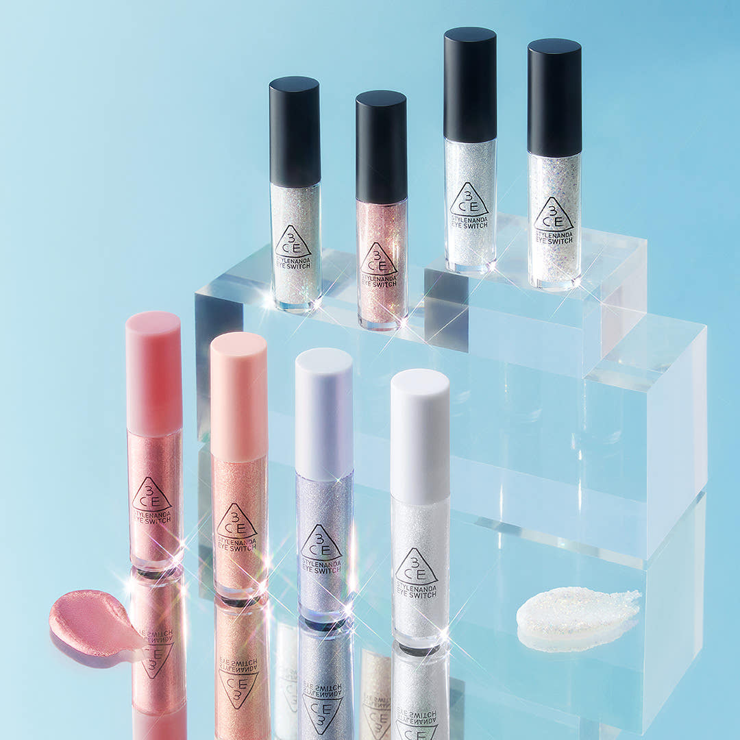 A collection of 3CE Eye Switch liquid eyeshadows in various shades with shimmer effect displayed on acrylic stands against a blue background.