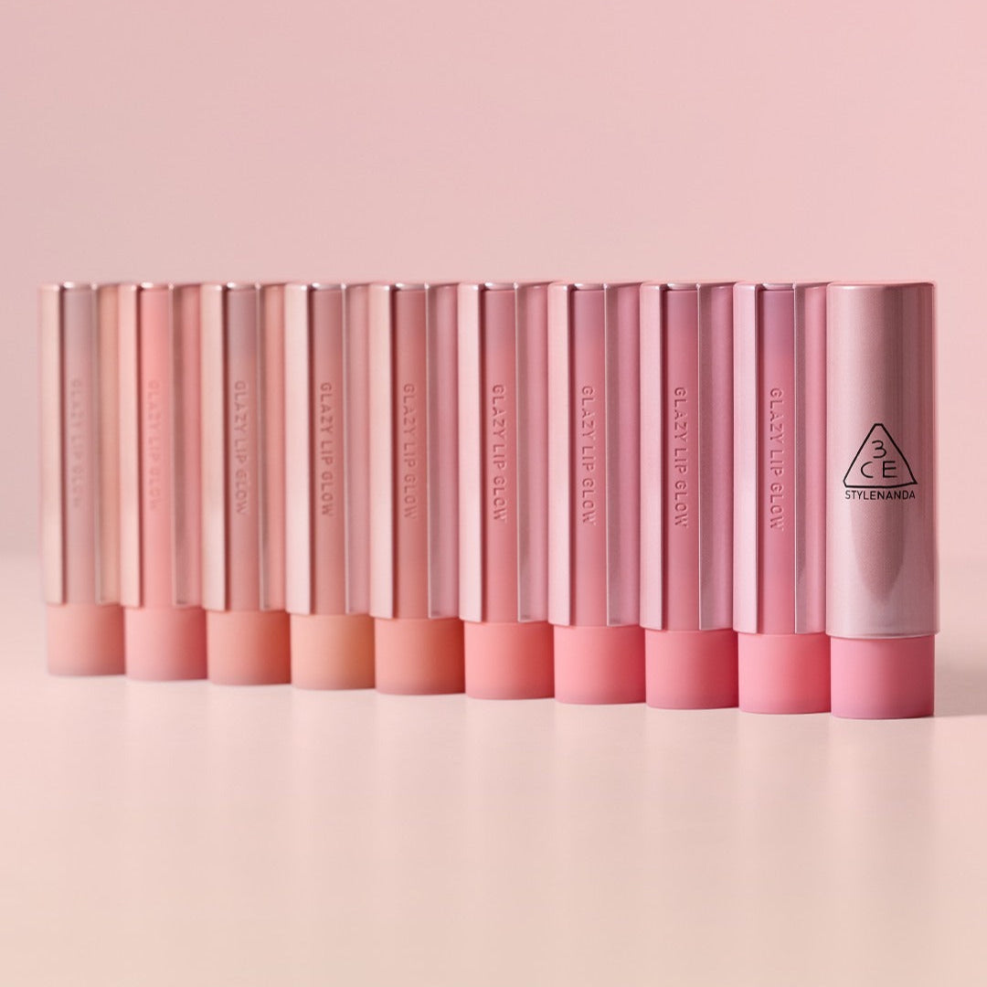 Assorted shades of 3CE Glaze Lip Glow 3.2g lipsticks lined up on a light pink background.