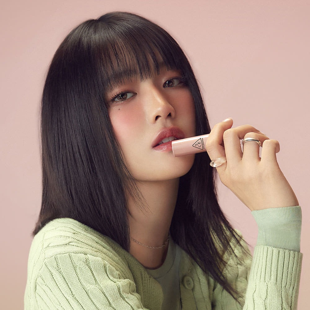Woman wearing soft makeup holding 3CE Glaze Lip Glow 3.2g against a pastel pink background.