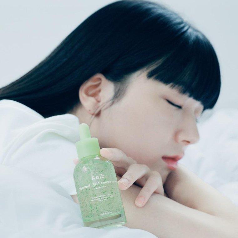 Woman resting with ABIB Heartleaf TECA Capsule Serum Calming Drop 50ml bottle, showcasing skincare product.