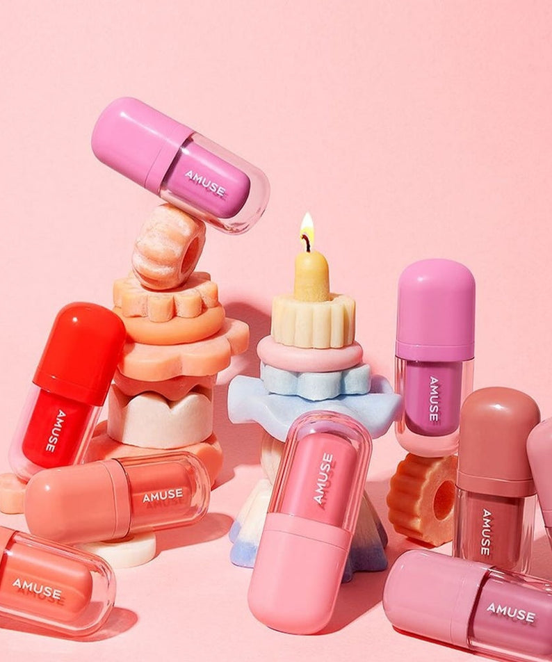 Colorful arrangement of AMUSE BEBE TINT lip tints with candle display against pink background.