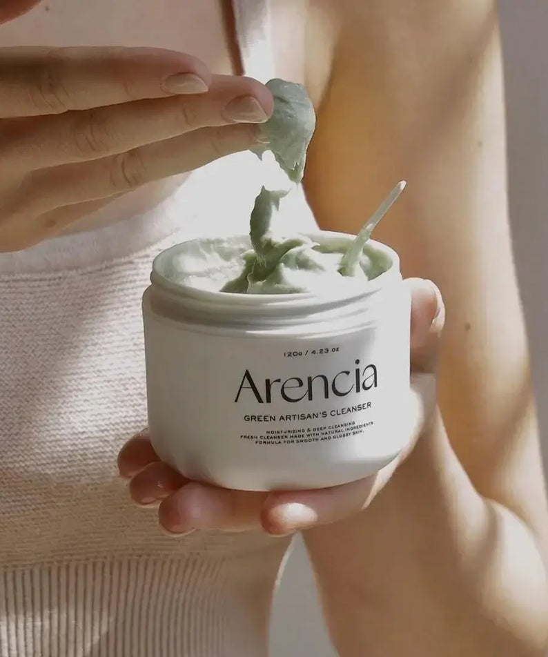 Person holding open jar of ARENCIA Green Artisan Skin Boosting Cleanser 120g with a spatula scooping product.