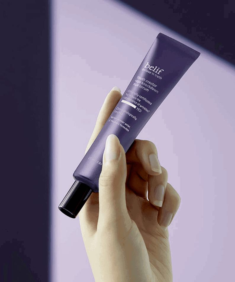 Hand holding BELIF Age Knockdown V Eye Serum 25ml against a purple background.