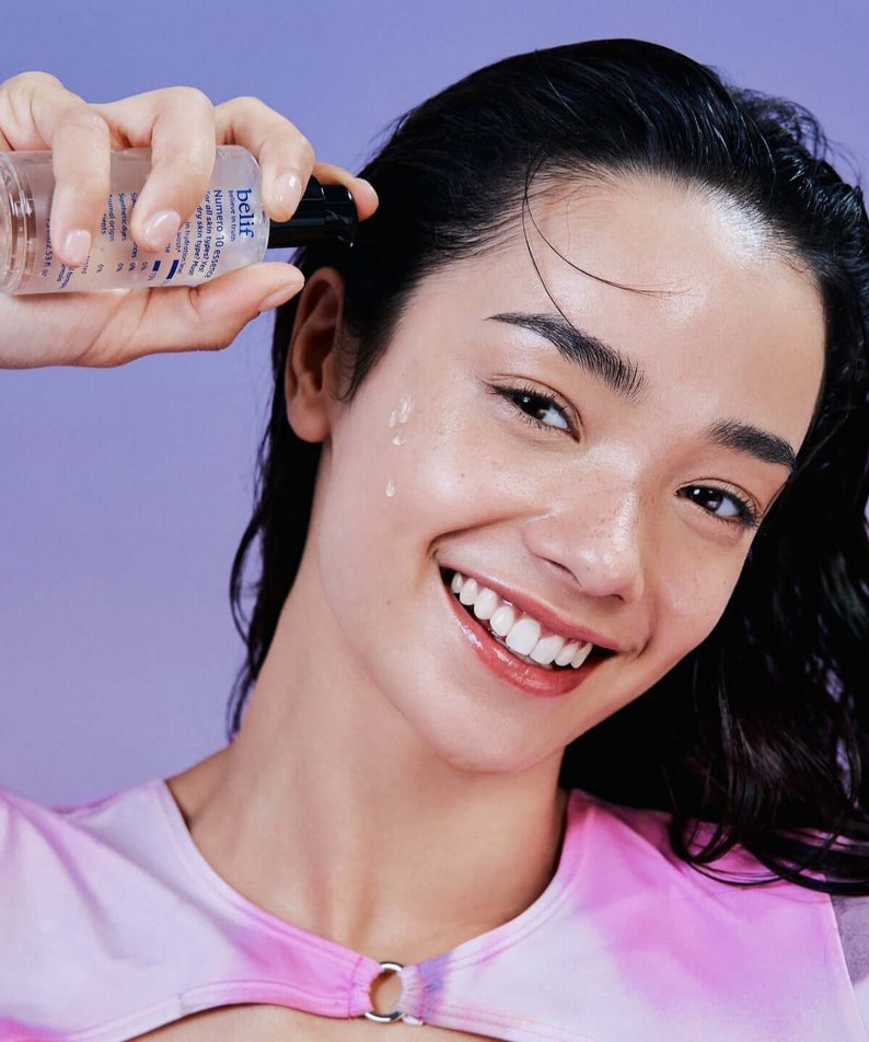 Woman applying BELIF Numero 10 Essence 75ml on her face, smiling brightly with a purple background.