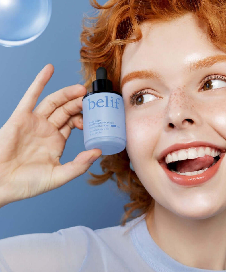Woman holding BELIF Super Drops Multi 7-Hyaluron Serum 30ml against a blue background, showcasing skincare product.