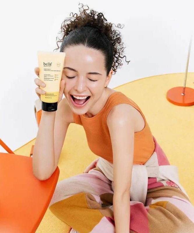Woman holding BELIF Super Knights bright Toning Vegan Pack Cleanser 150ml, smiling on a colorful background