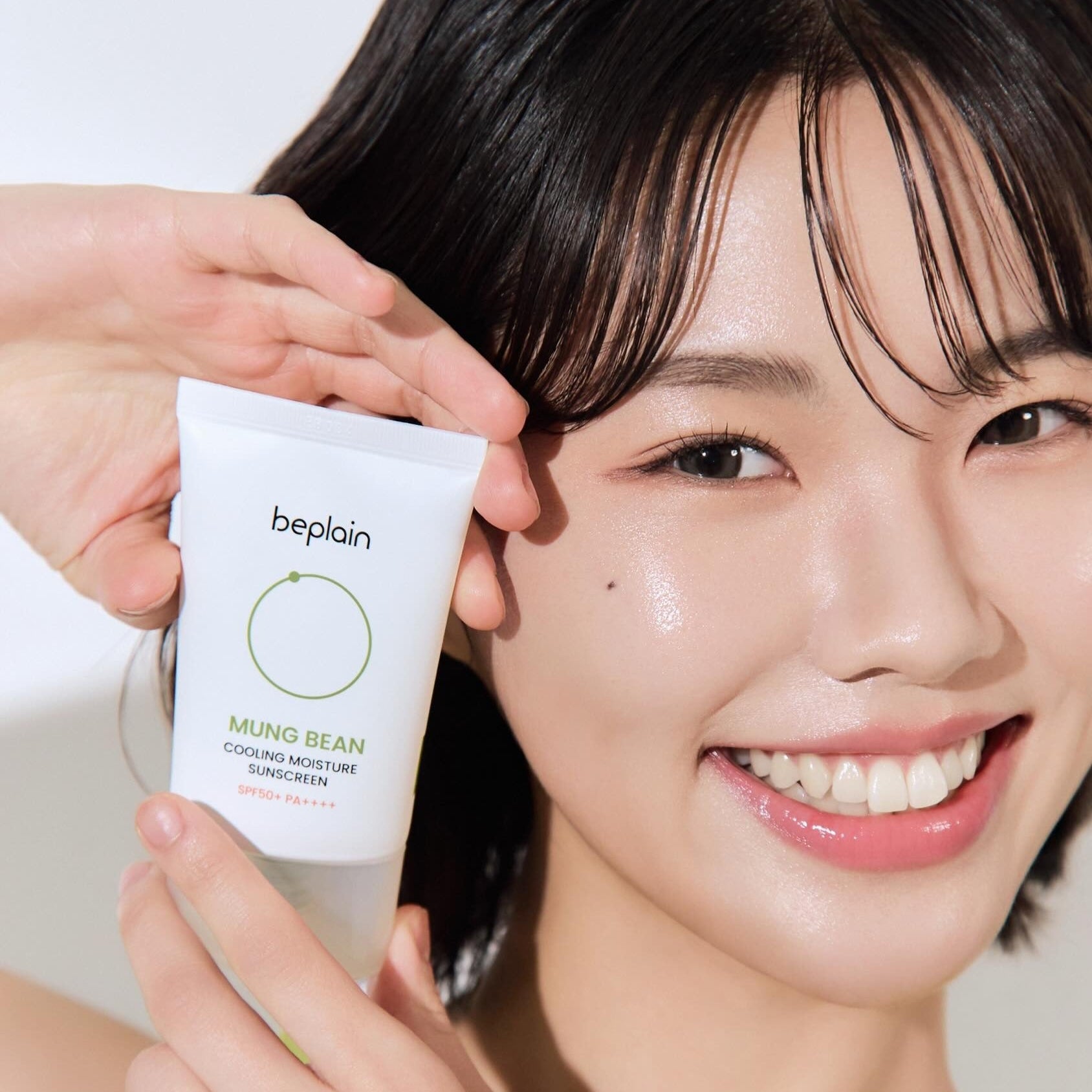 Smiling woman holding BEPLAIN Green Bean Cooling Moisture Sun Cream 50ml tube next to her face.