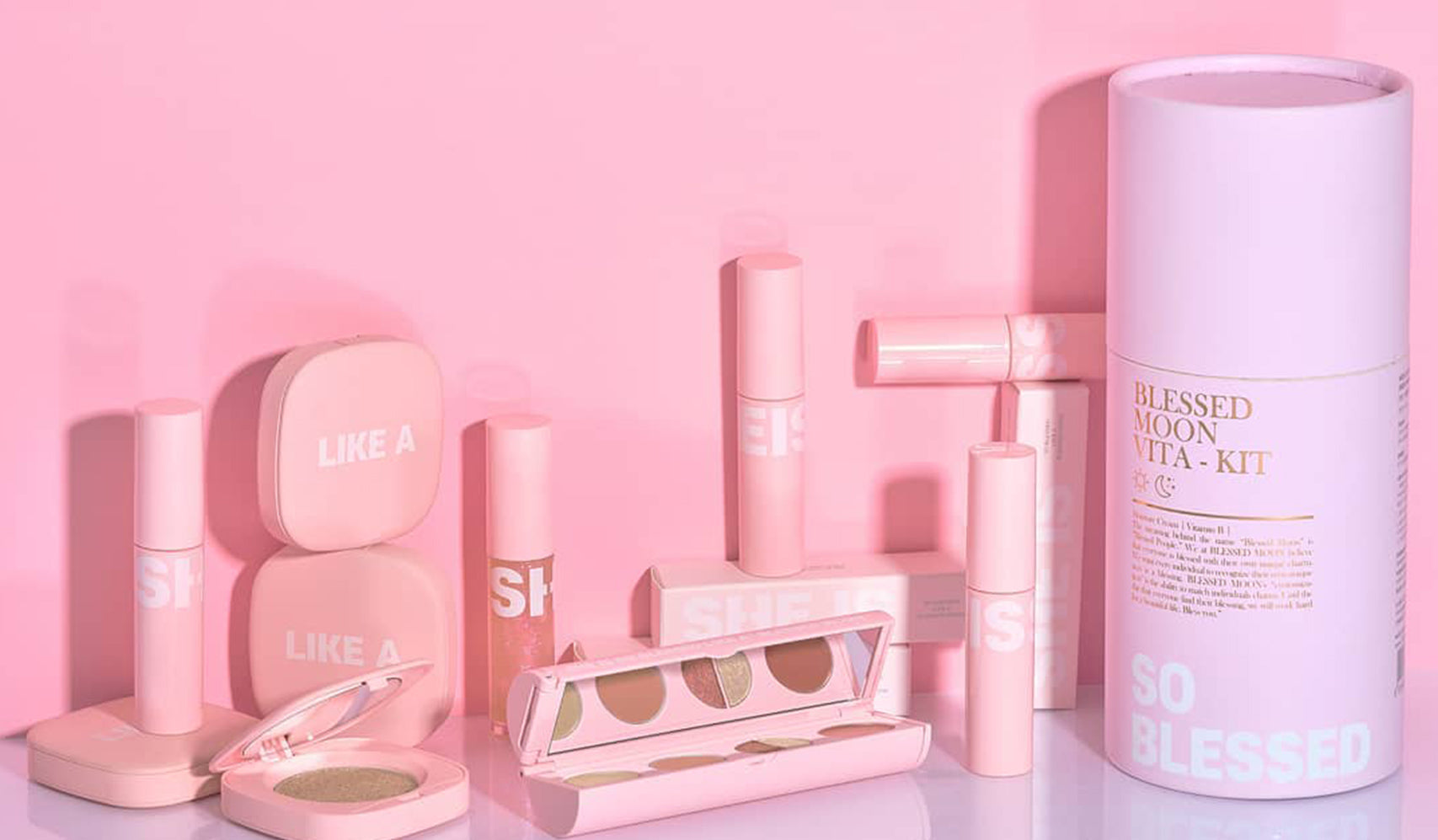 Assorted pink makeup products including lipsticks, blush, eyeshadow palette on a pink background.