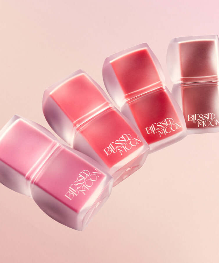 BLESSED MOON Veil Blur Lip Tint in various shades on a pink background, four lip tints lined up with sleek packaging.