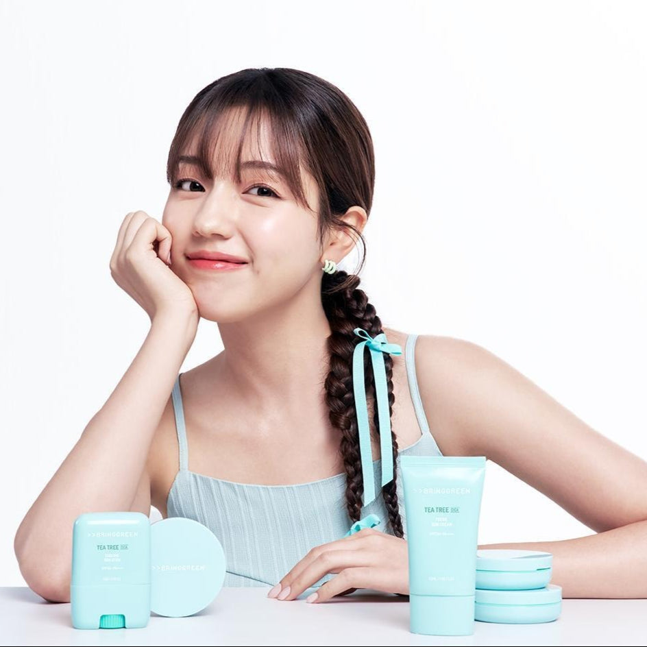 Smiling woman with BRINGGREEN Tea Tree Cica Tone Up Sun Cushion and skincare products on table.