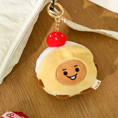 BT21 Bakery Plushy Keyring attaché à une chaîne dorée, posé sur une table en bois, design adorable de personnage patisserie.