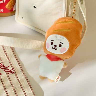 BT21 Bakery Plushy Keyring en forme de viennoiserie, accroché à un sac en toile, illustration de style adorable et kawaii en tissu doux.