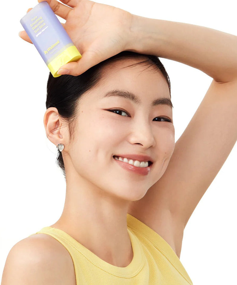 Woman smiling and holding BY WISHTREND Pore Smoothing Bakuchoil Sun Stick 18g, showcasing skincare product.
