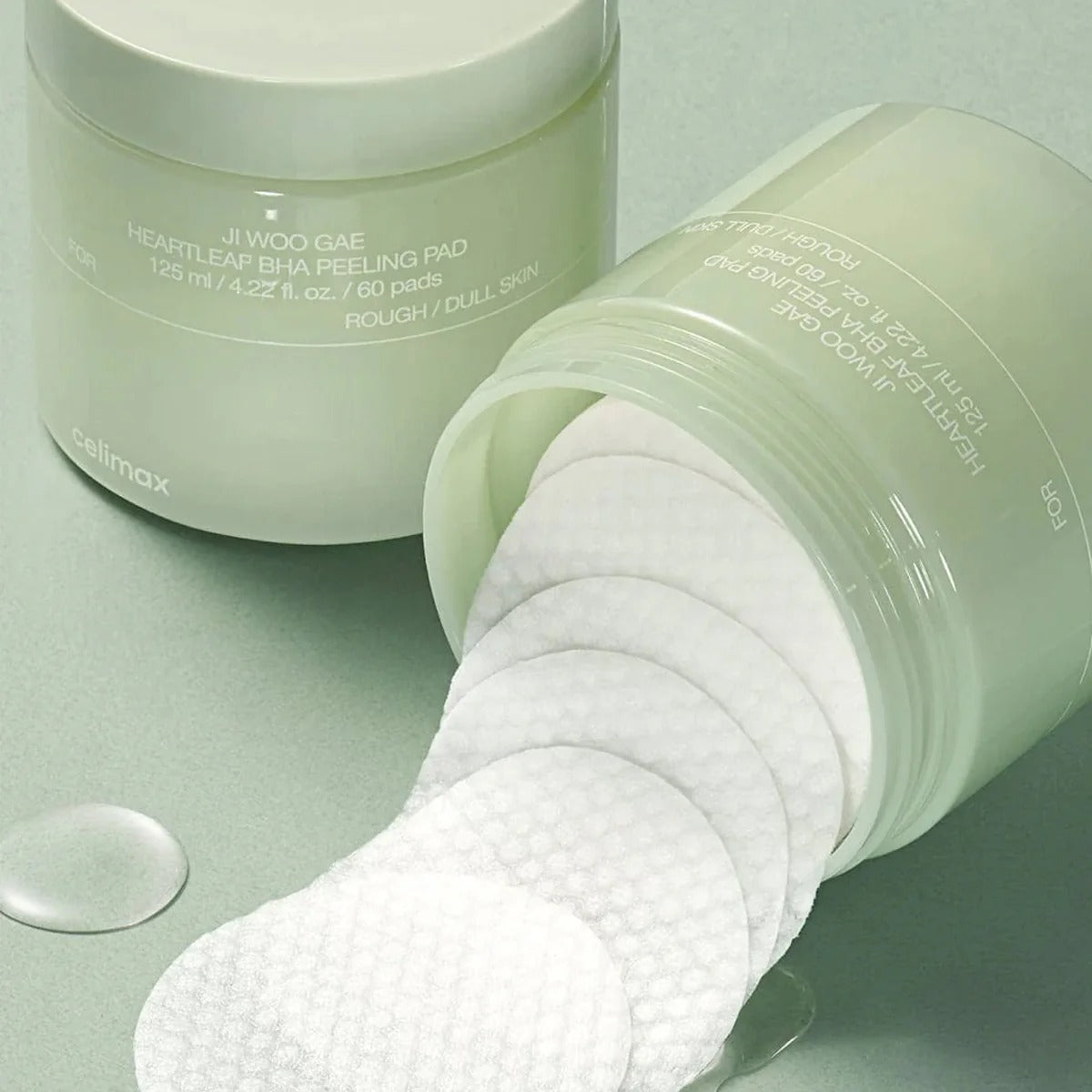 Open jar of CELIMAX Ji Woo Gae Heartleaf BHA Peeling Pad 60 Pads on green background, showcasing texture and packaging.